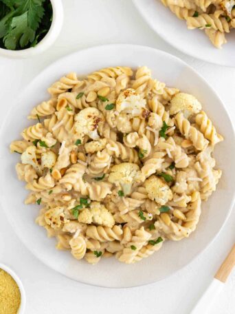 roasted cauliflower alfredo pasta on two ceramic plates beside a bowl of pine nuts and parsley