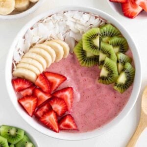 a strawberry kiwi smoothie bowl surrounded by bowls of sliced banana, strawberries, and kiwi fruit