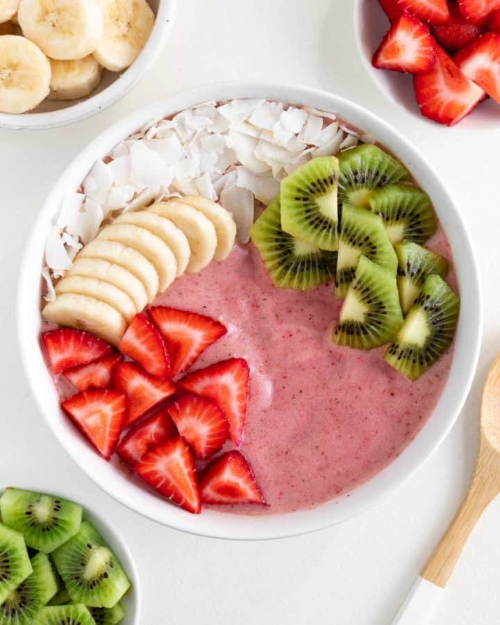 a strawberry kiwi smoothie bowl surrounded by bowls of sliced banana, strawberries, and kiwi fruit