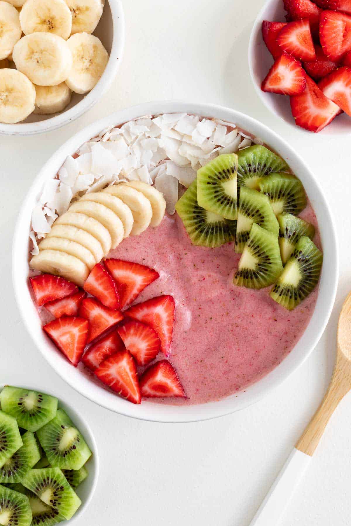 a strawberry kiwi smoothie bowl surrounded by bowls of sliced banana, strawberries, and kiwi fruit