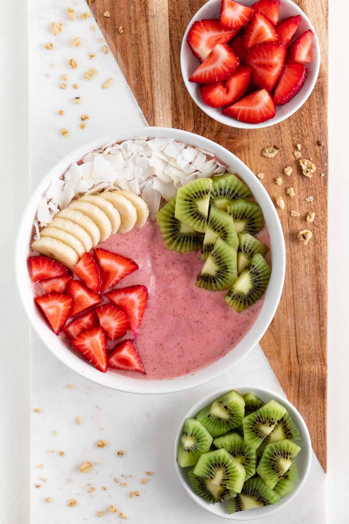 a marble and wood board topped with a strawberry kiwi smoothie bowl, granola, a bowl of berries, and a bowl of kiwi fruit