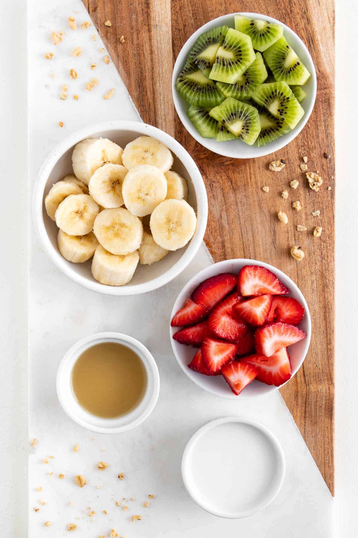 a marble and wood cutting board topped with bowls of banana, kiwi fruit, berries, lime juice, and almond milk