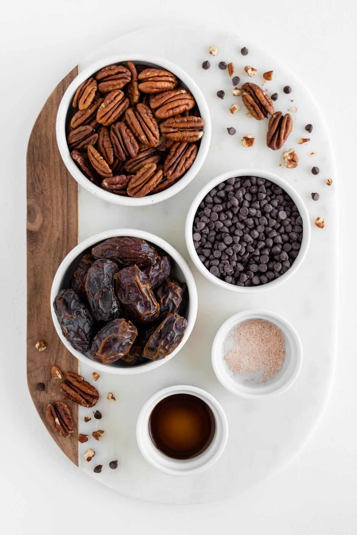 a marble board with bowls of raw pecans, medjool dates, chocolate chips, salt, and vanilla extract