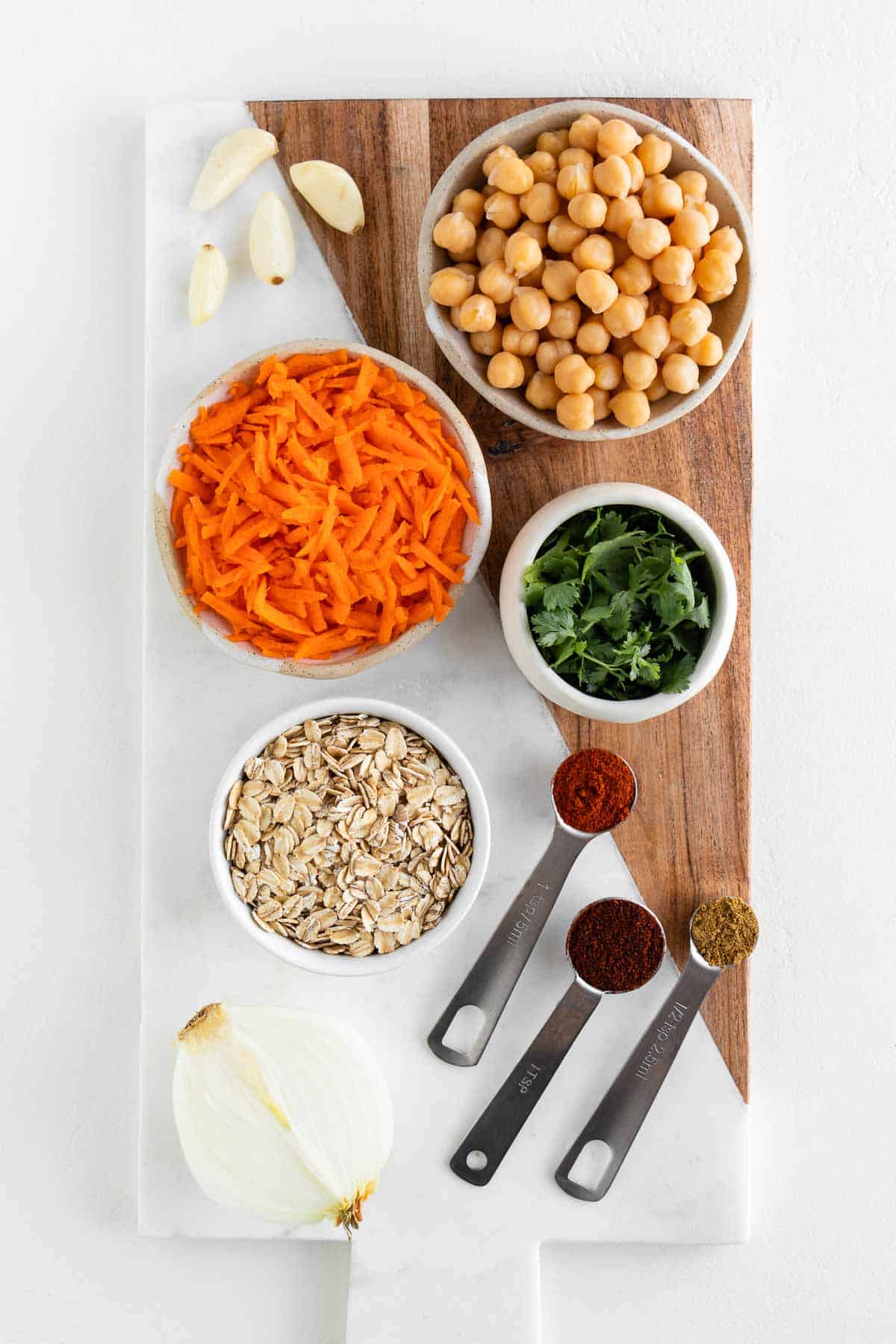 a marble and wooden cutting board topped with chickpeas, grated carrots, cilantro, rolled oats, onion, garlic, and spices