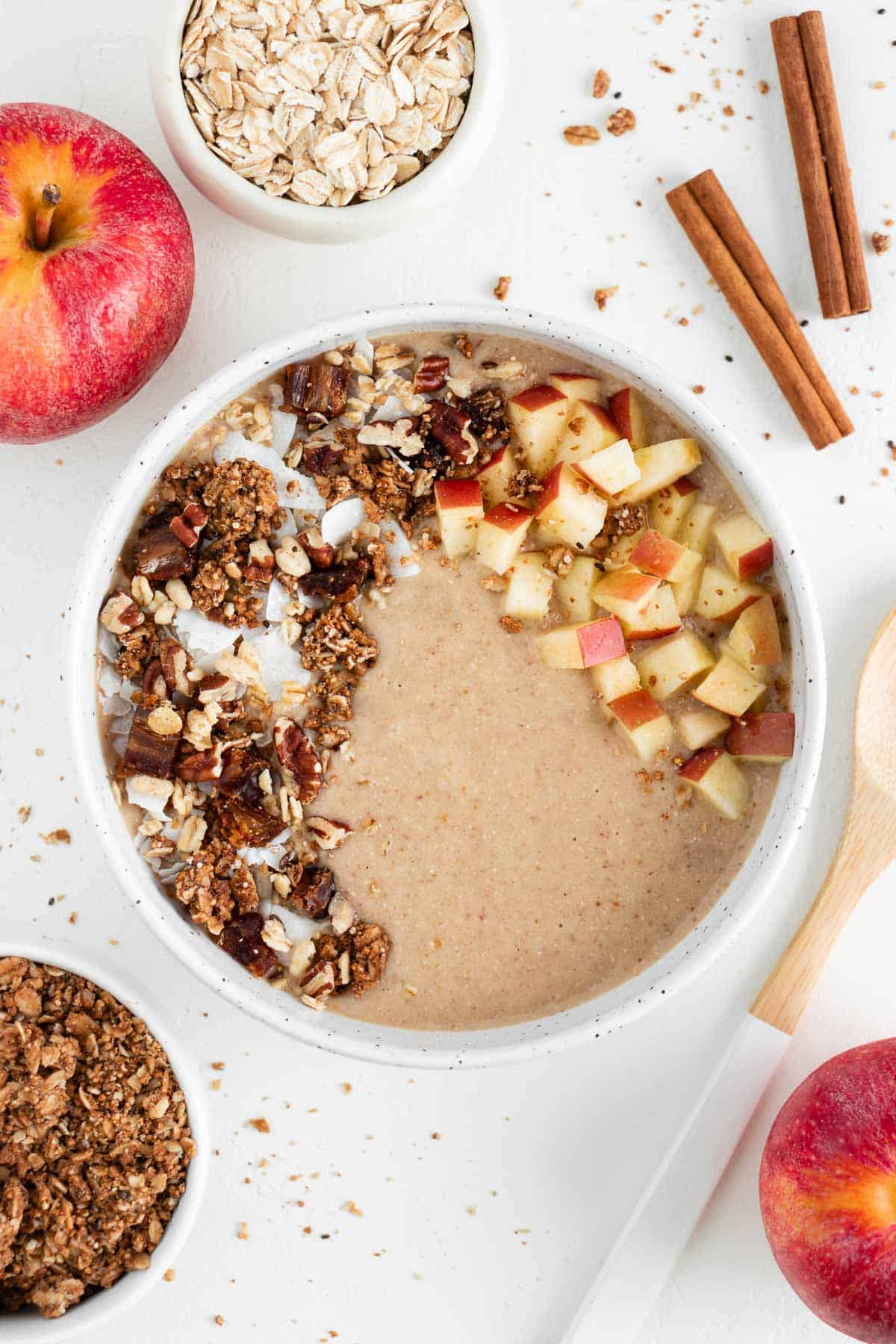 an apple pie smoothie bowl in a ceramic bowl surrounded by apples, oats, granola, and cinnamon sticks