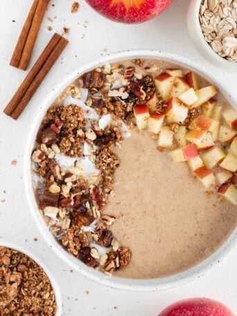 an apple pie smoothie bowl inside a ceramic bowl surrounded by oats, granola, cinnamon sticks, and apples