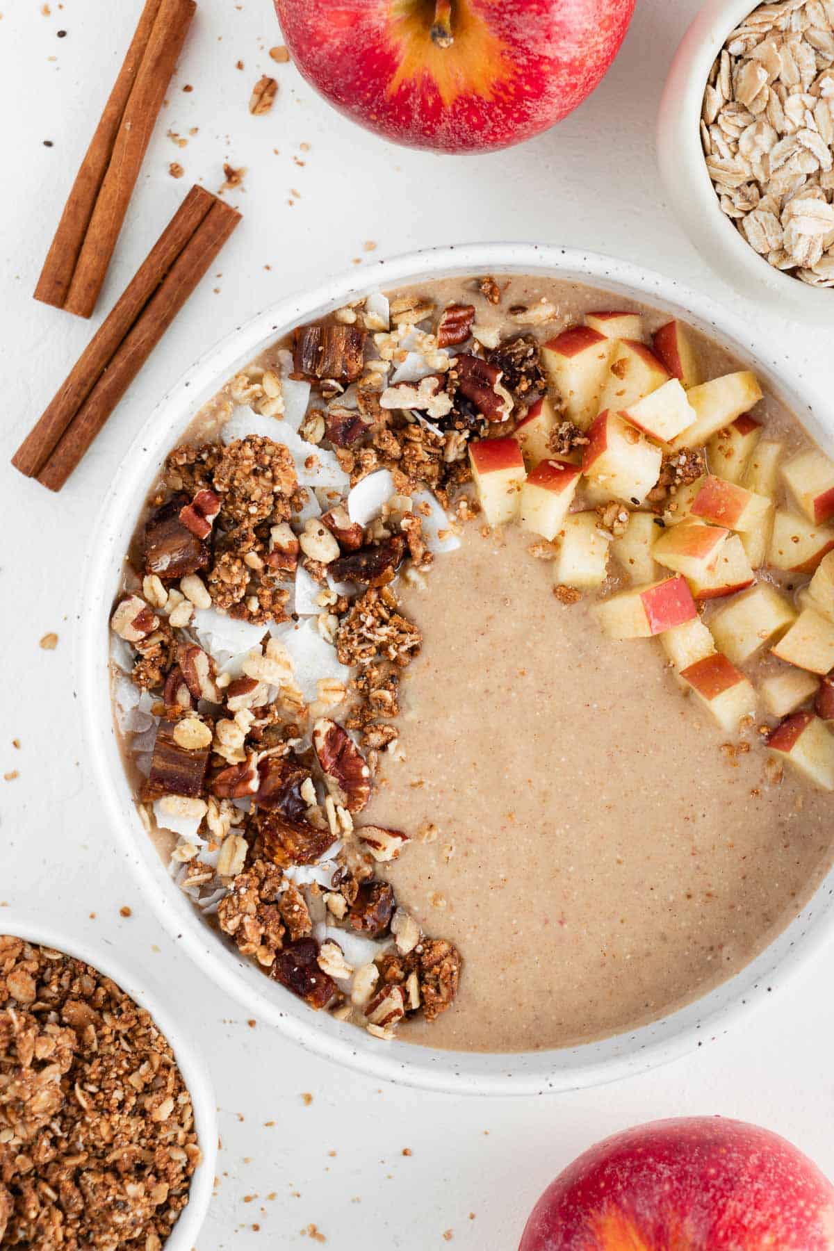 an apple pie smoothie bowl inside a ceramic bowl surrounded by oats, granola, cinnamon sticks, and apples