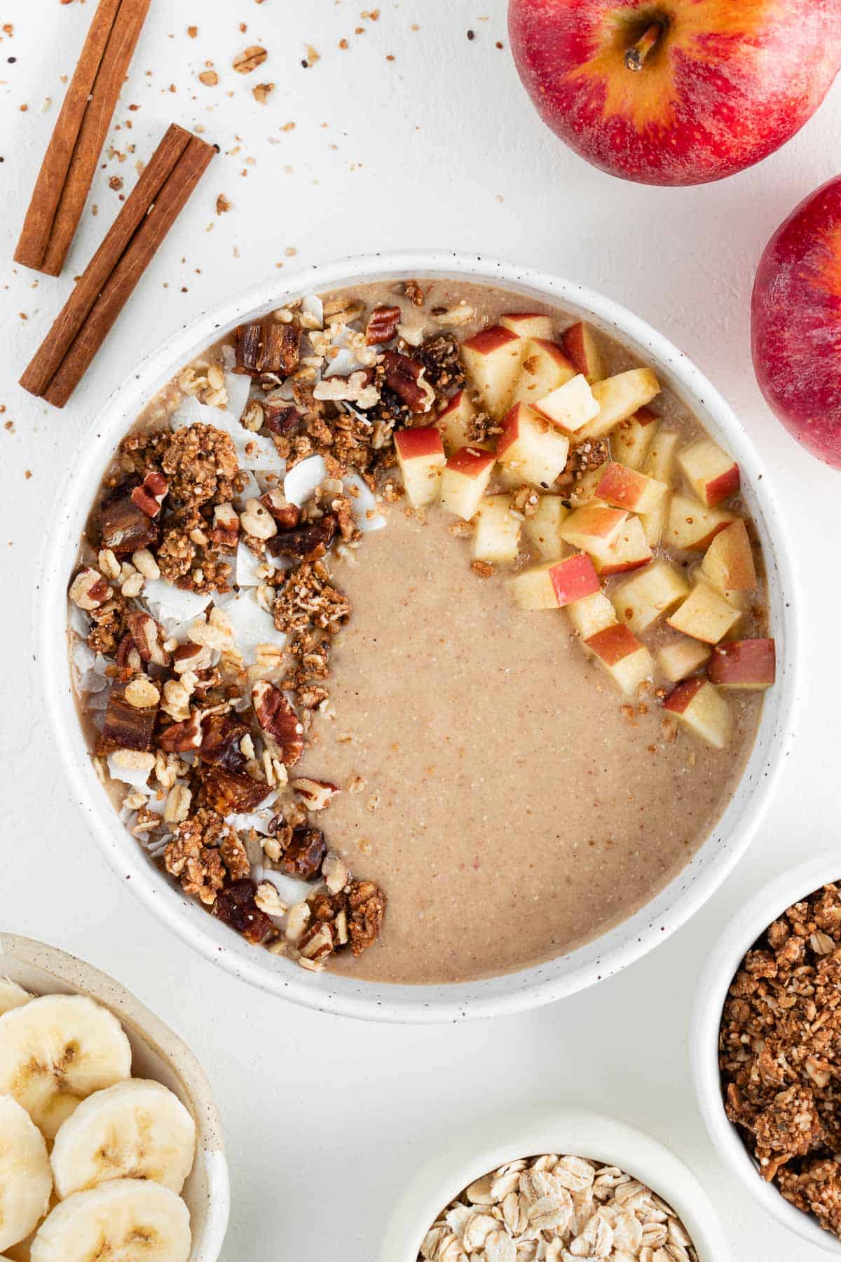 apple pie smoothie bowl surrounded by cinnamons ticks, apples, sliced banana, oats, and granola