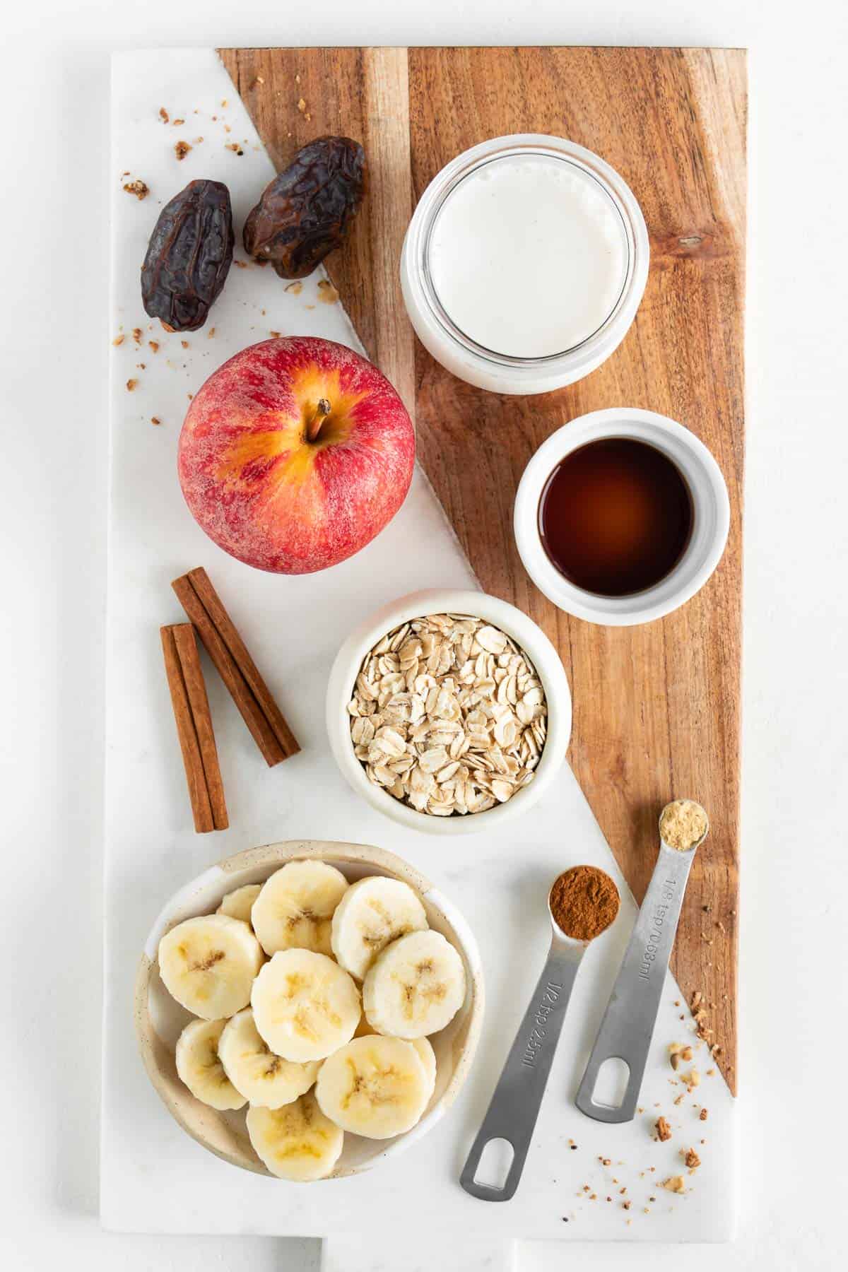 a marble board topped with an apple, oats, sliced banana, oat milk, medjool dates, cinnamon sticks, and vanilla extract