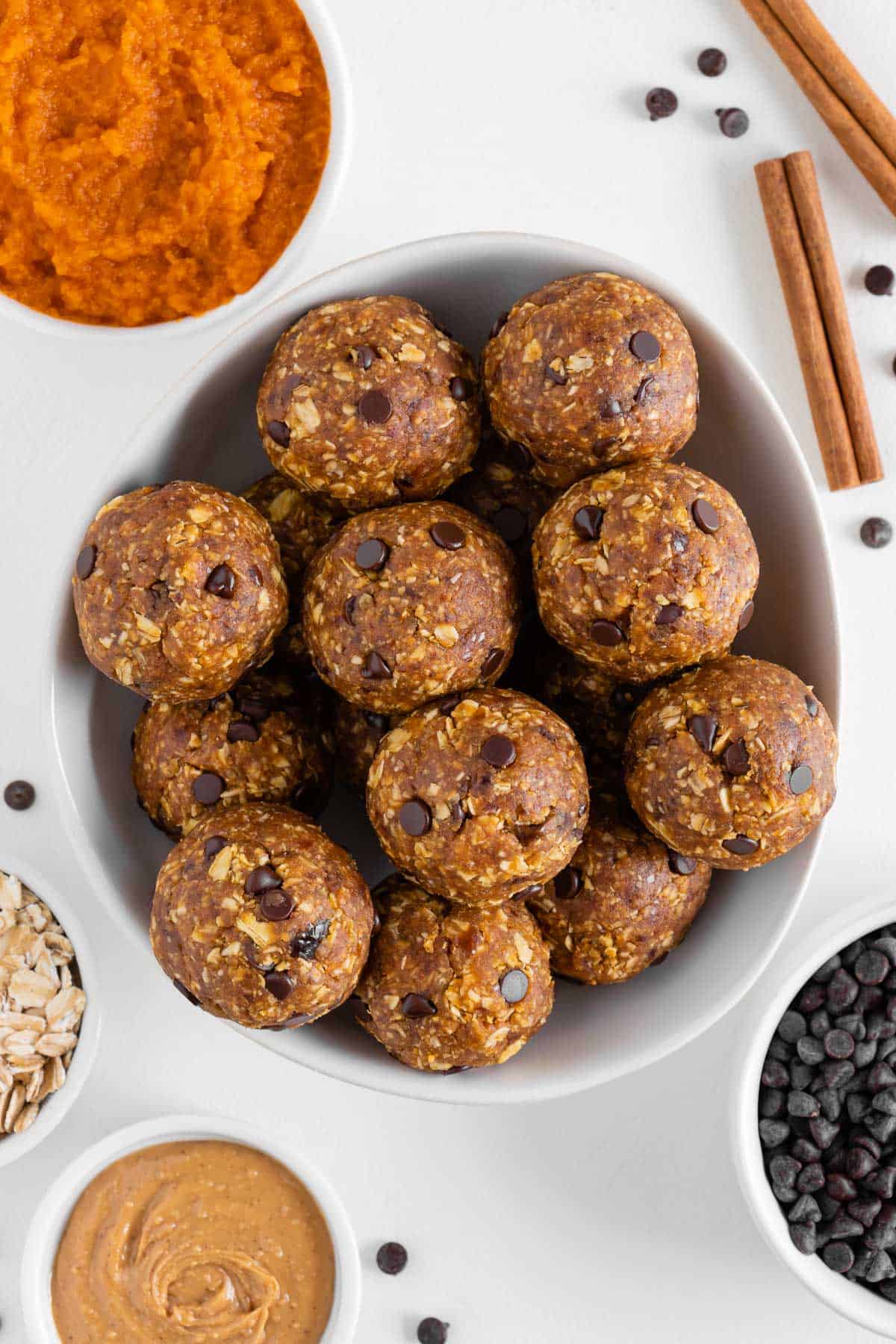 pumpkin energy balls in a white bowl surrounded by pumpkin puree, peanut butter, chocolate chips, and oats