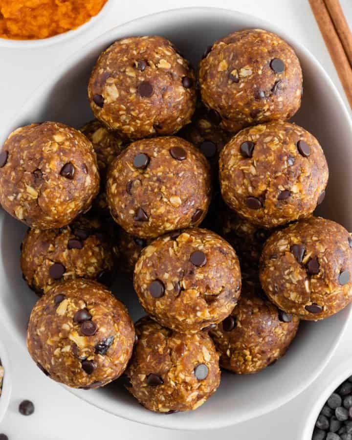pumpkin energy balls in a white bowl surrounded by pumpkin puree, peanut butter, chocolate chips, and oats