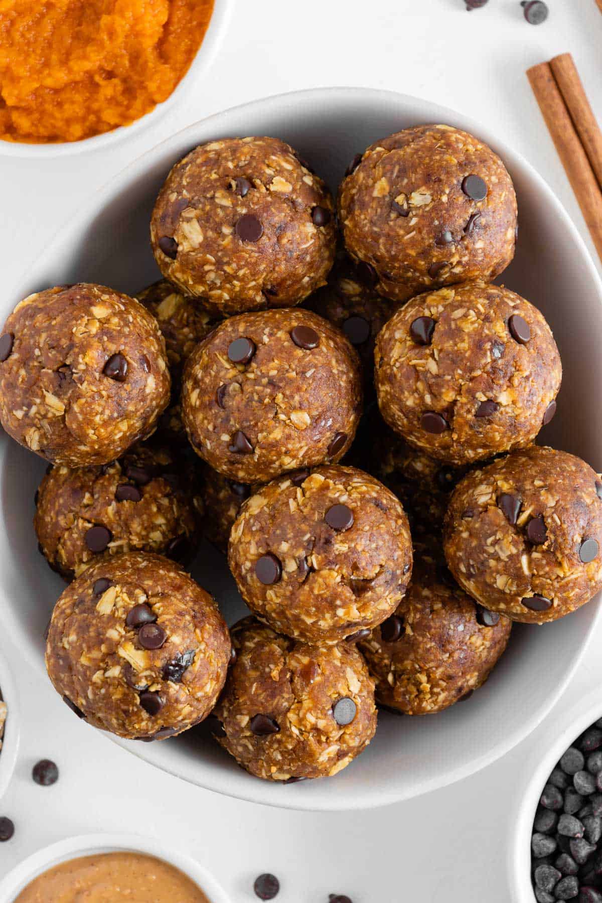 pumpkin energy balls in a white bowl surrounded by pumpkin puree, peanut butter, chocolate chips, and oats