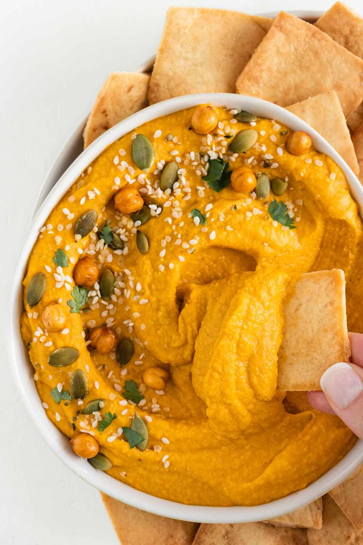a hand holding a pita chip scooping pumpkin hummus out of a bowl