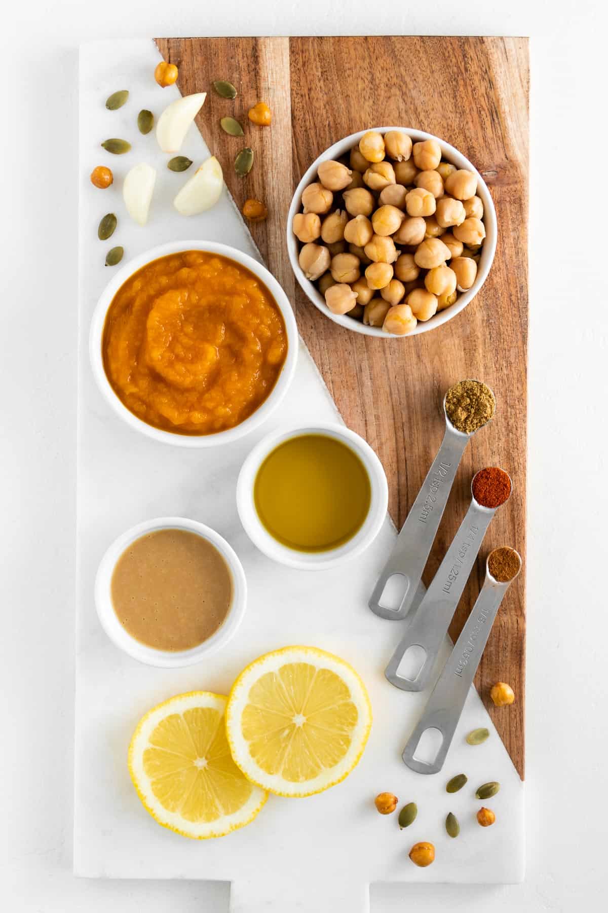 a marble and wood board topped with bowls of chickpeas, pumpkin puree, olive oil, tahini, sliced lemon, cumin, paprika, and cinnamon