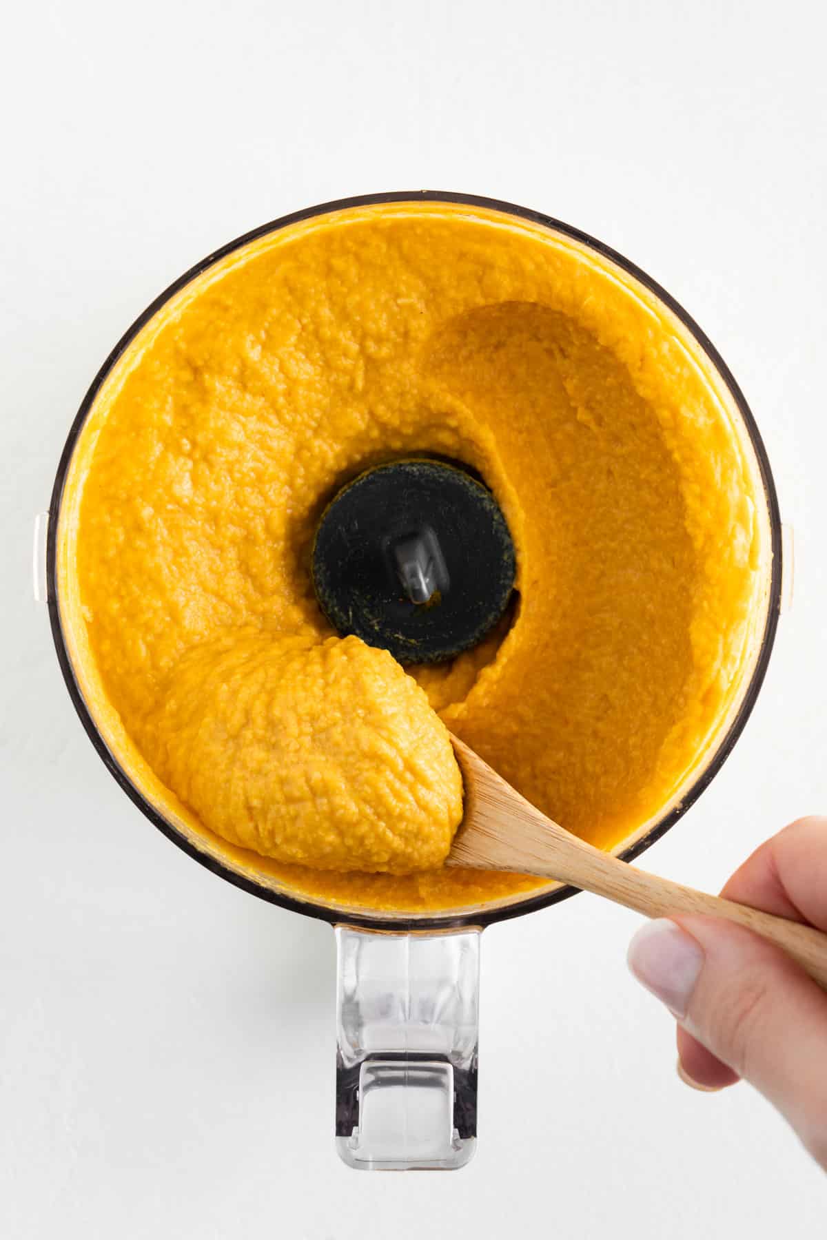 a hand holding a wooden spoon scooping pumpkin hummus inside the bowl of a food processor