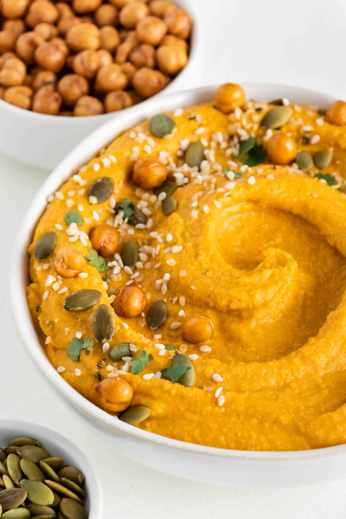 pumpkin hummus in a white bowl topped with chickpeas, pumpkin seeds, and sesame seeds