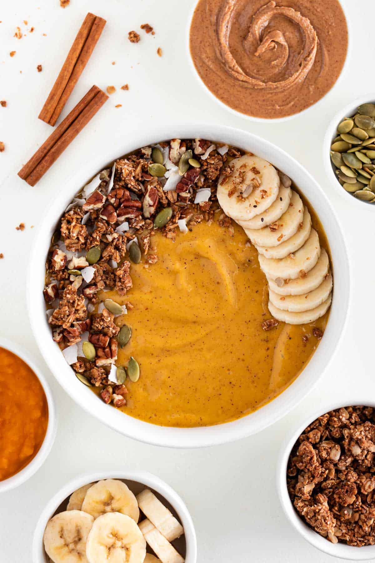 pumpkin pie smoothie bowl inside a white bowl surrounded by almond butter, pumpkin, banana, seeds, and granola