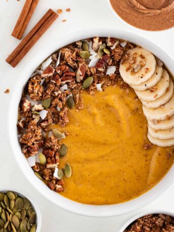 a pumpkin pie smoothie inside a white bowl topped with granola, banana, pumpkin seeds, and coconut flakes