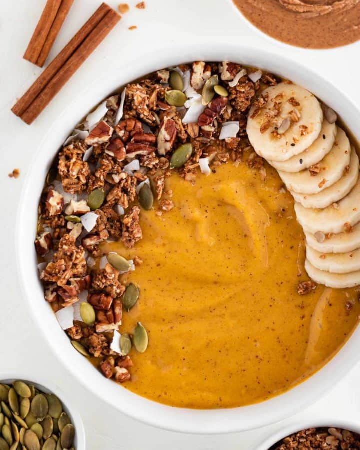 a pumpkin pie smoothie inside a white bowl topped with granola, banana, pumpkin seeds, and coconut flakes