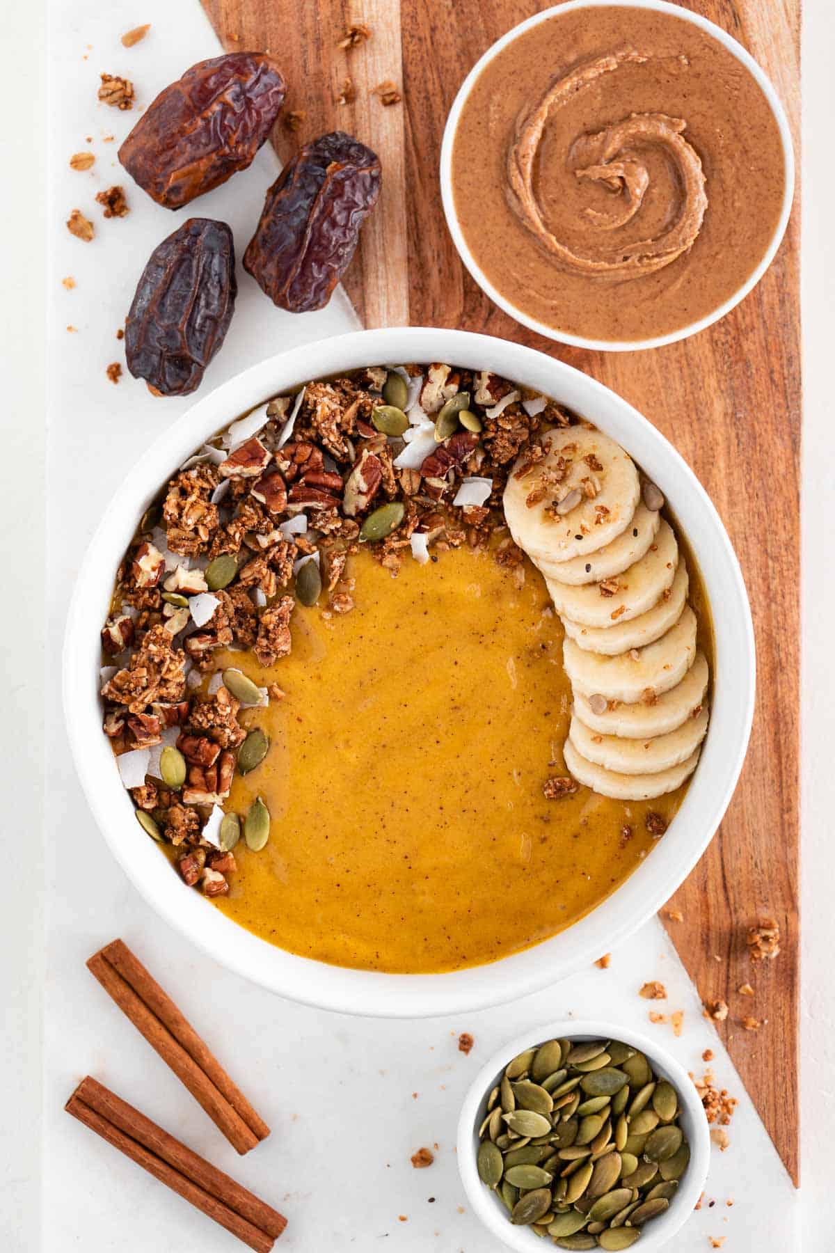 a pumpkin pie smoothie bowl on top of a marble cutting board along with medjool dates, almond butter, and pumpkin seeds