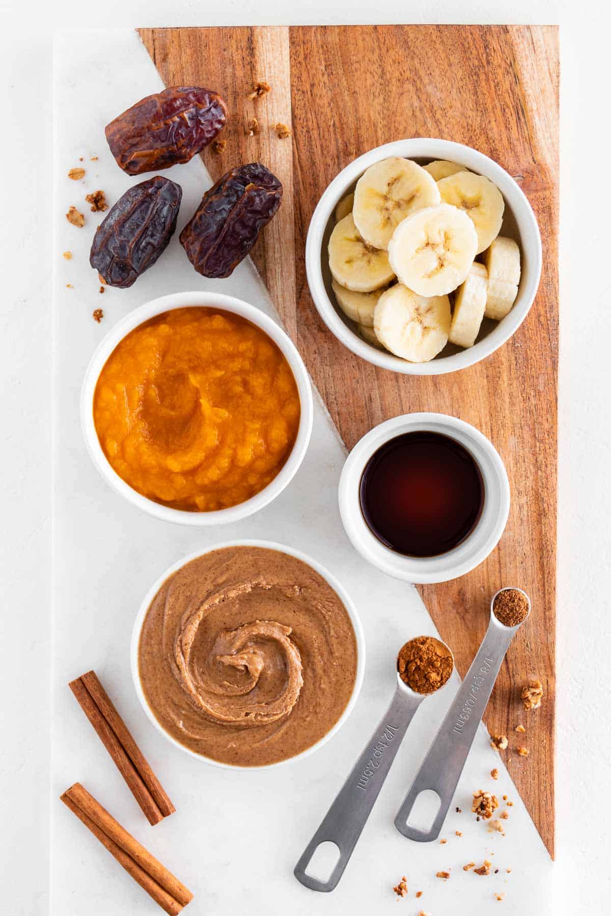 bowls of pumpkin puree, almond butter, sliced banana, medjool dates, vanilla extract, cinnamon sticks, and granola on a marble cutting board
