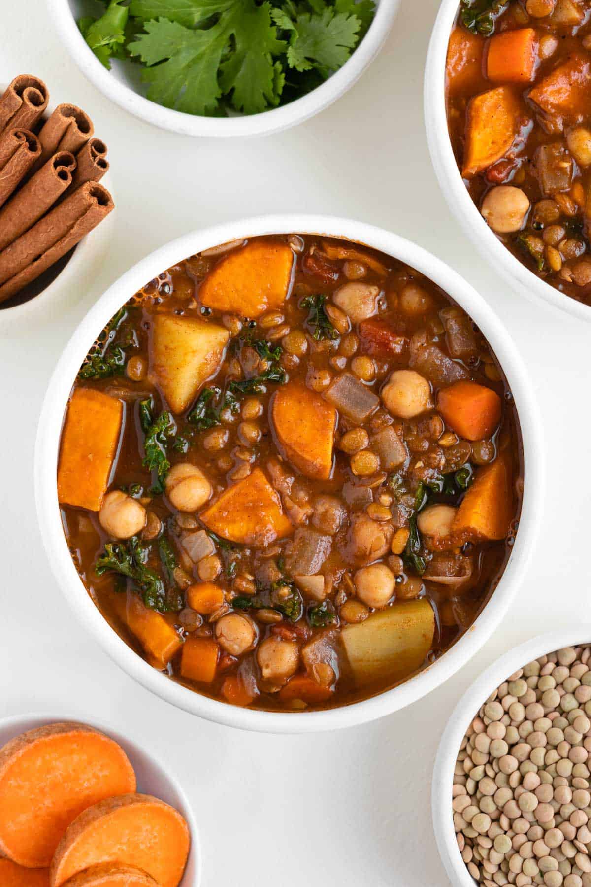 vegan moroccan stew inside two bowls beside fresh cilantro, green lentils, cinnamon, and sliced sweet potato