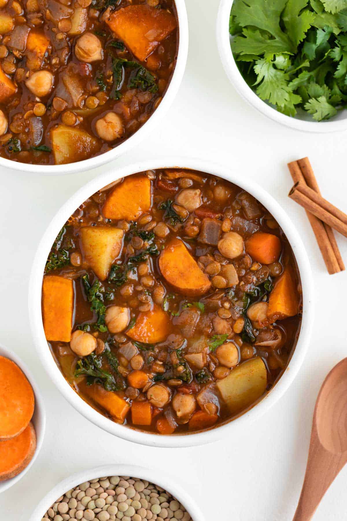 vegan moroccan stew inside two bowls beside fresh cilantro, green lentils, cinnamon, and sliced sweet potato