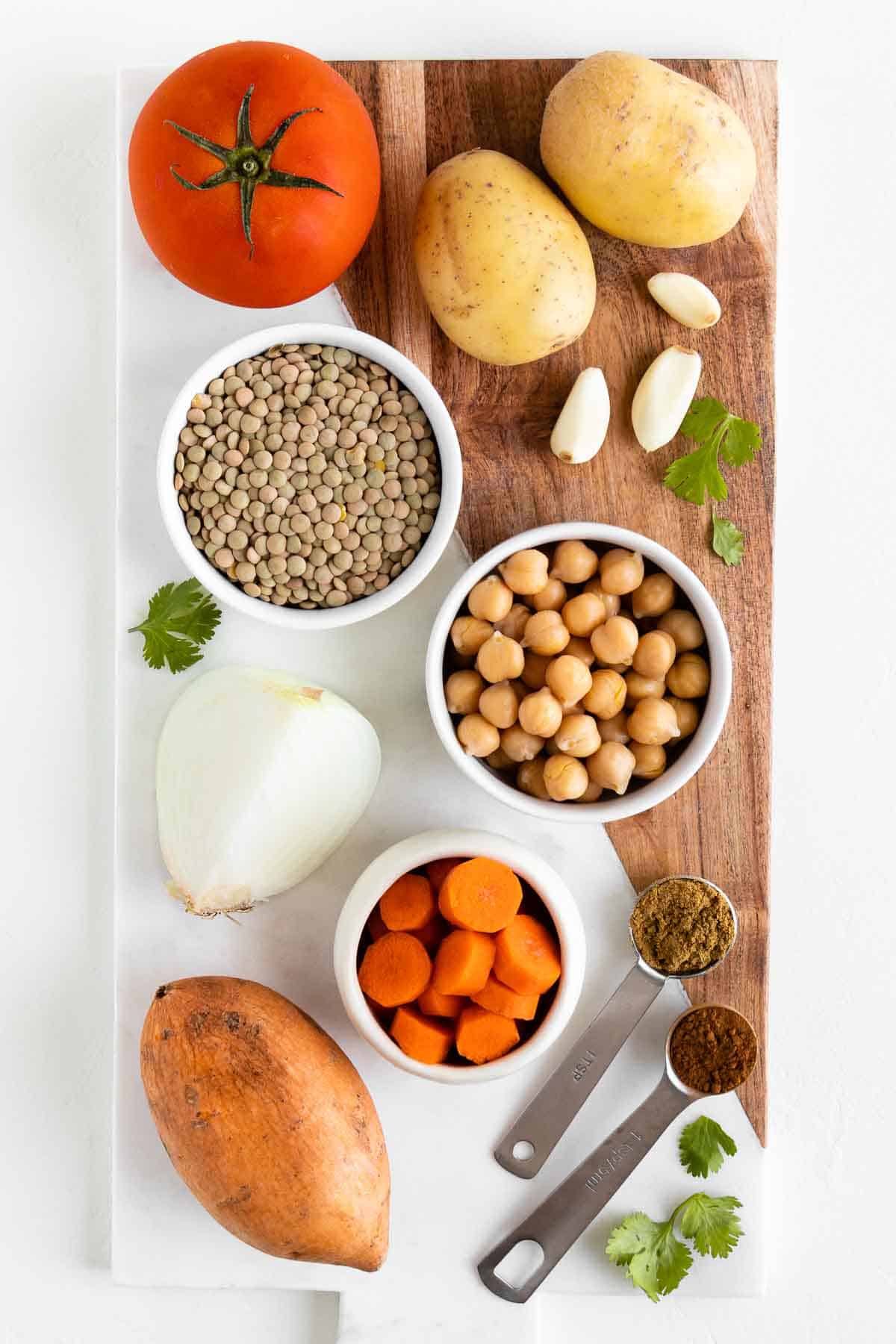 a small bowl of green lentils, chickpeas, and carrots on a marble board along with sweet potatoes, gold potatoes, tomato, and spices