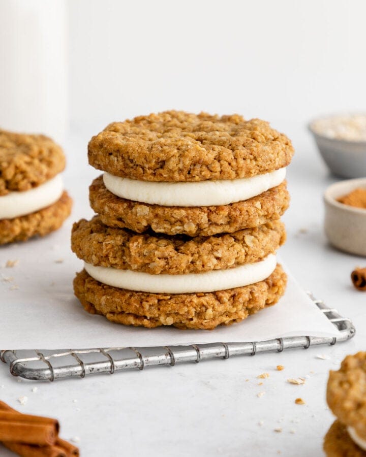 a stack of three vegan oatmeal cream pie cookies beside a jar of almond milk, a bowl of oats, and cinnamon