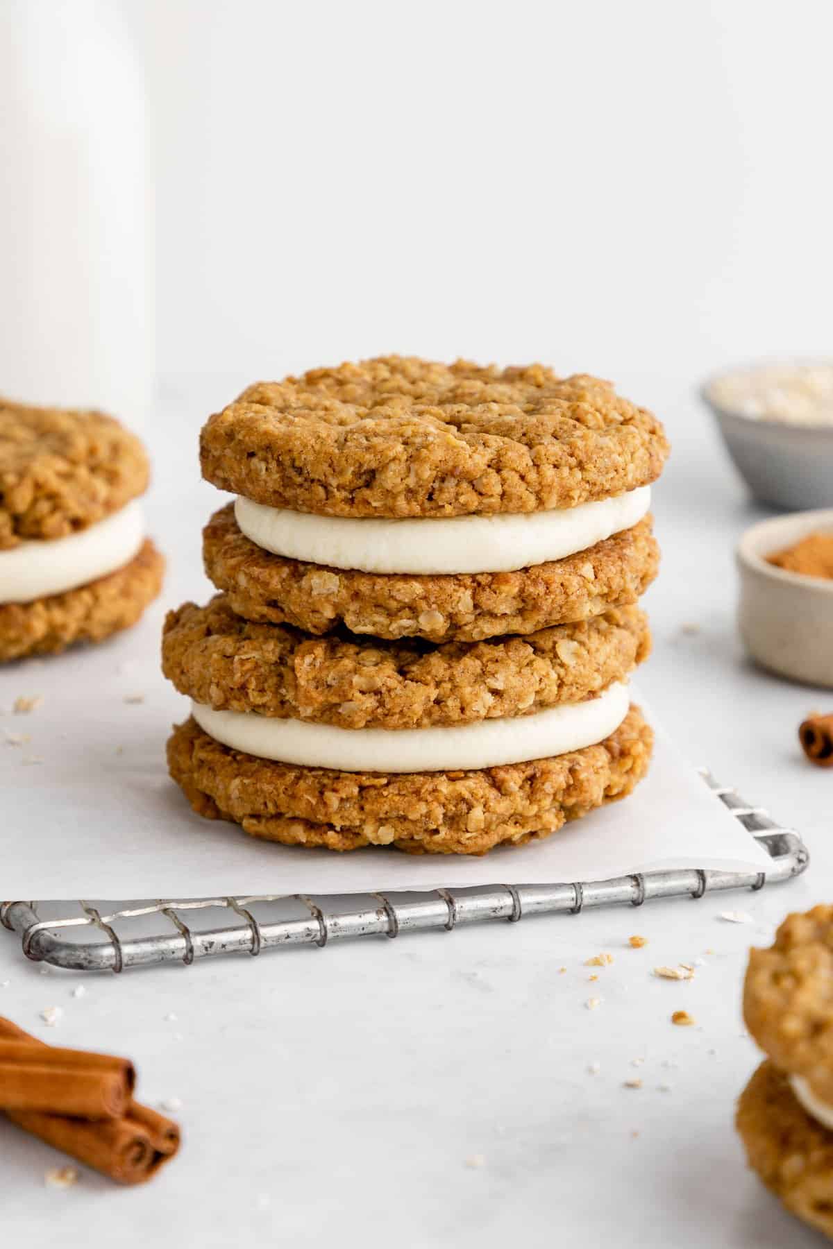 a stack of three vegan oatmeal cream pie cookies beside a jar of almond milk, a bowl of oats, and cinnamon