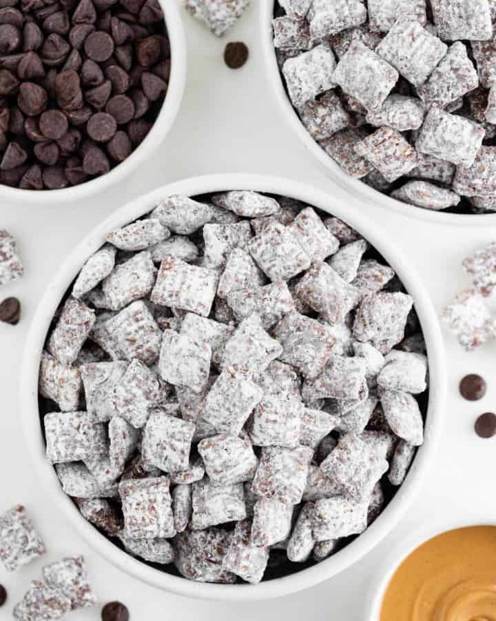 two bowls of vegan puppy chow surrounded by a bowl of chocolate chips and peanut butter