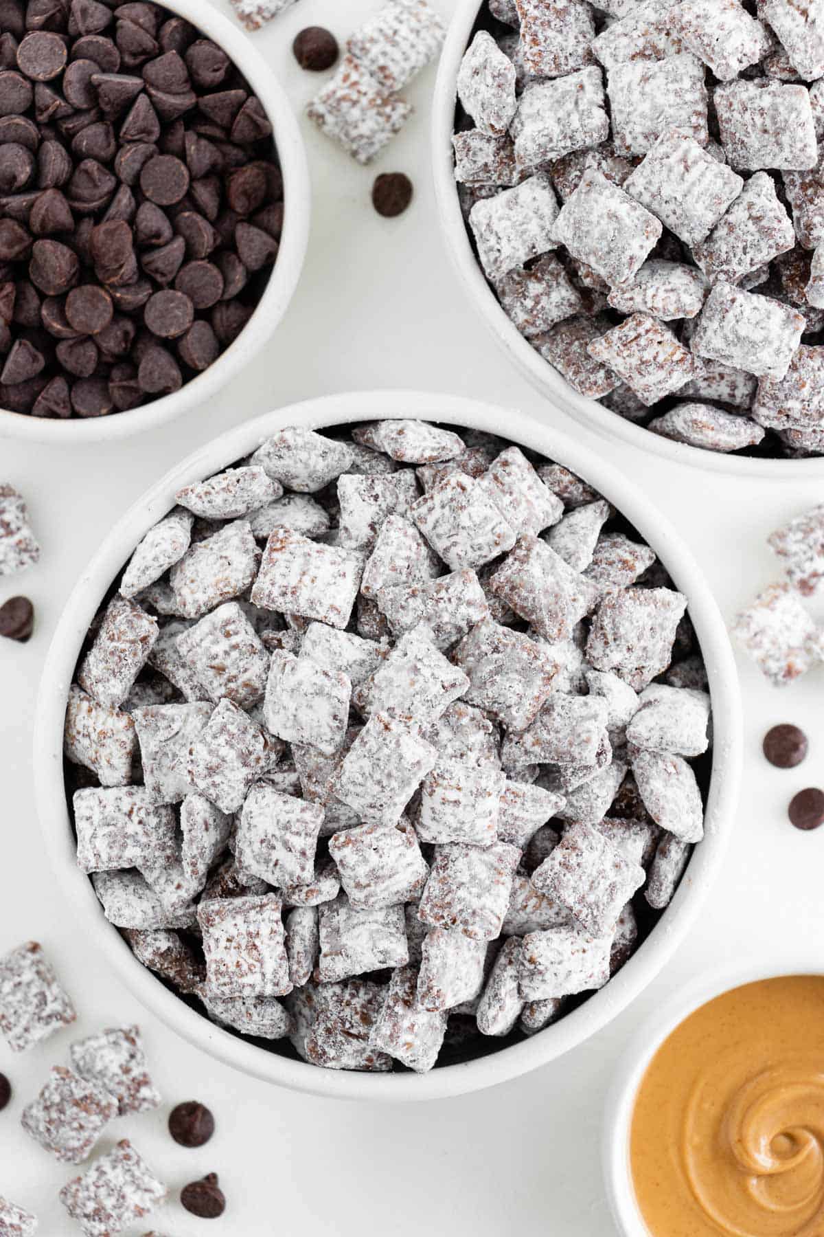 two bowls of vegan puppy chow surrounded by a bowl of chocolate chips and peanut butter