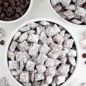 two bowls of vegan puppy chow surrounded by a bowl of chocolate chips and powdered sugar