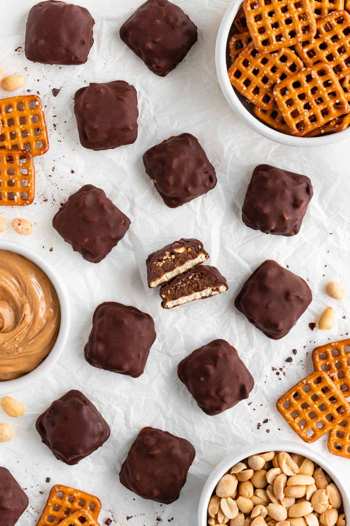 vegan homemade take 5 candy bars on white parchment paper surrounded by a bowl of peanut butter, a bowl of snap pretzels, and a bowl of peanuts