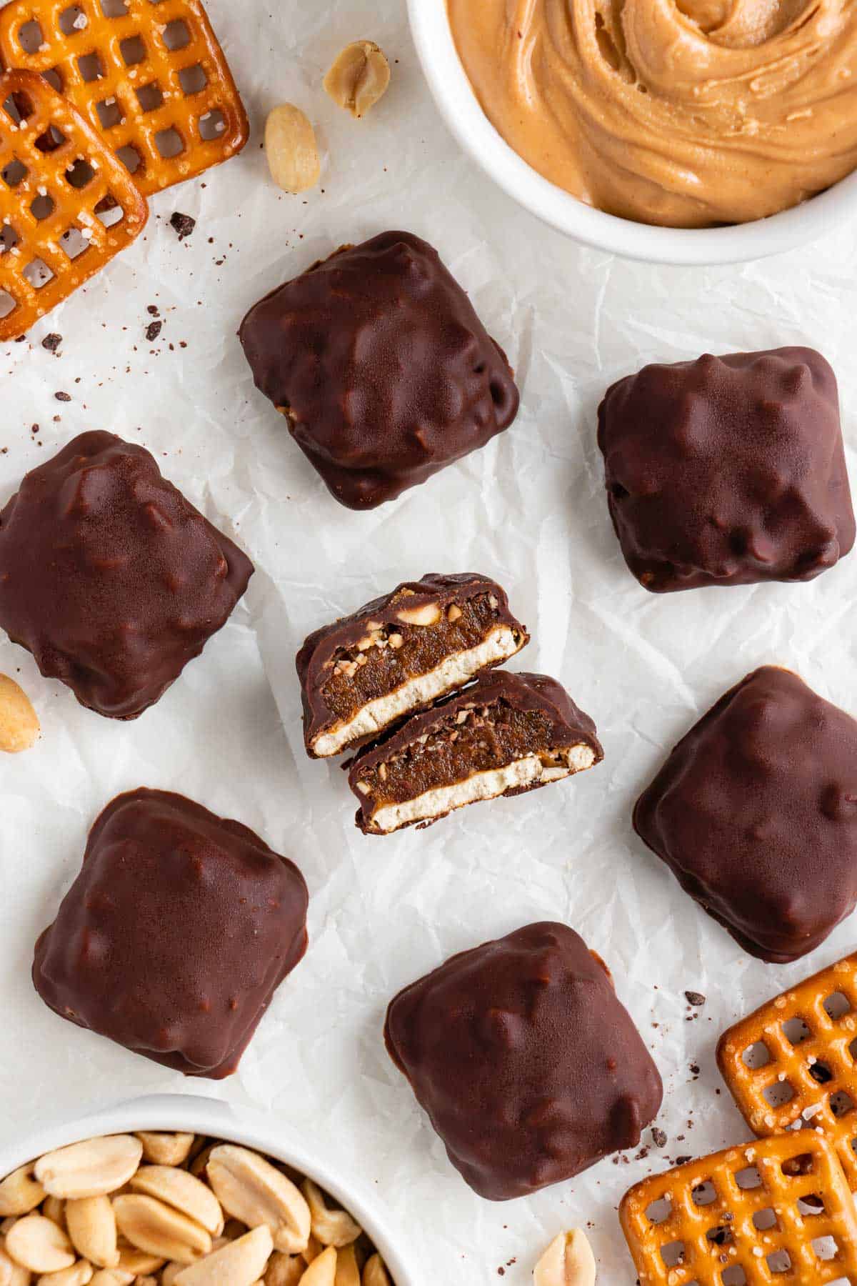 vegan homemade take 5 candy bars on white parchment paper surrounded by a bowl of peanut butter, a bowl of peanuts, and snap pretzels