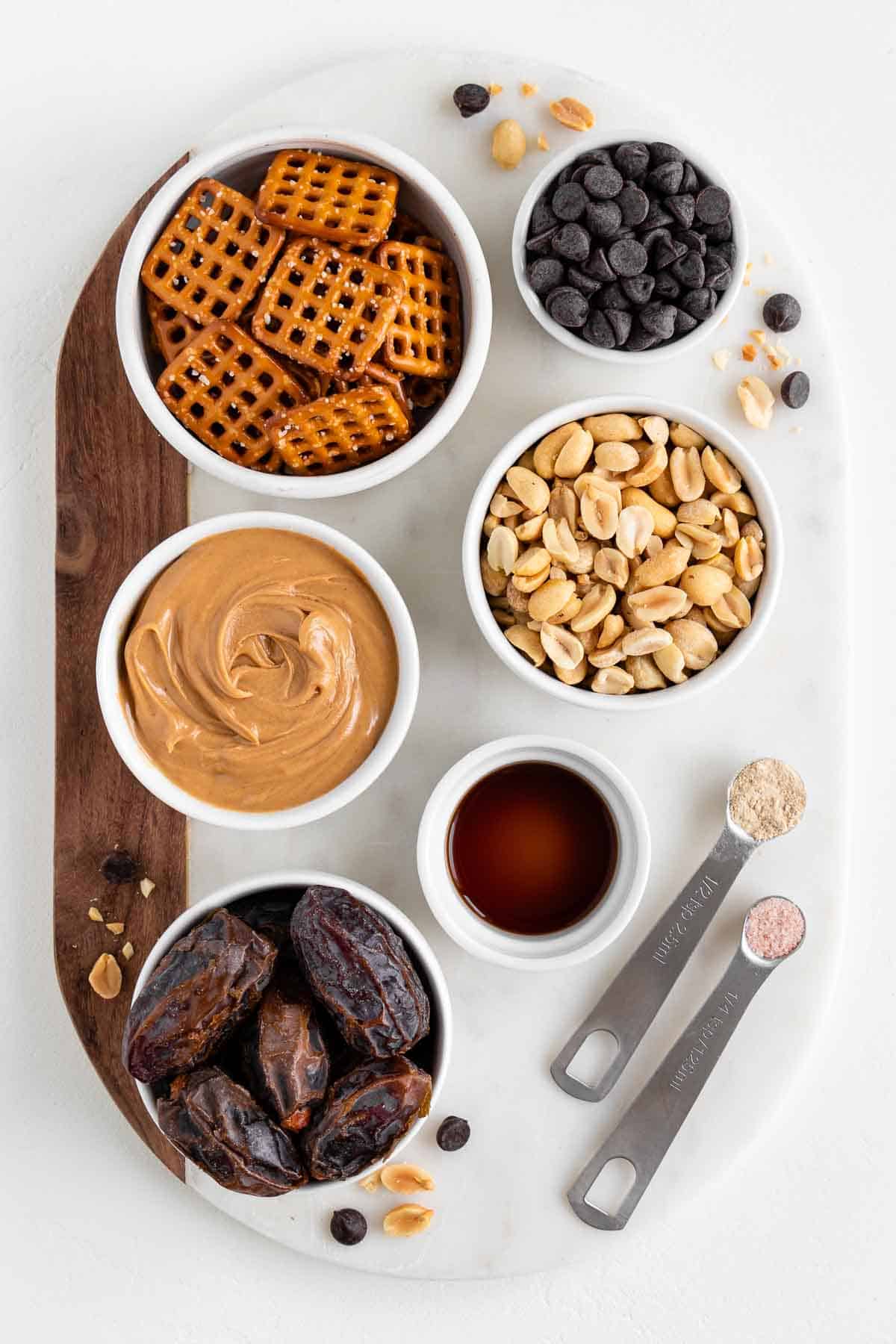 a round marble board topped with small white bowls of pretzels, peanut butter, peanuts, medjool dates, chocolate chips, and vanilla extract