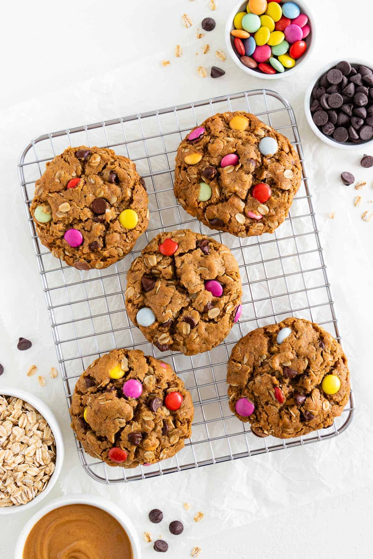 five vegan monster cookies on a small metal cookie rack beside bowls of oatmeal, chocolate chips, m&ms, and peanut butter