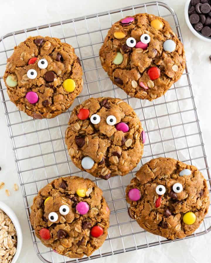five googly eye vegan monster cookies on a small metal cookie rack beside bowls of oatmeal, chocolate chips, m&ms, and peanut butter