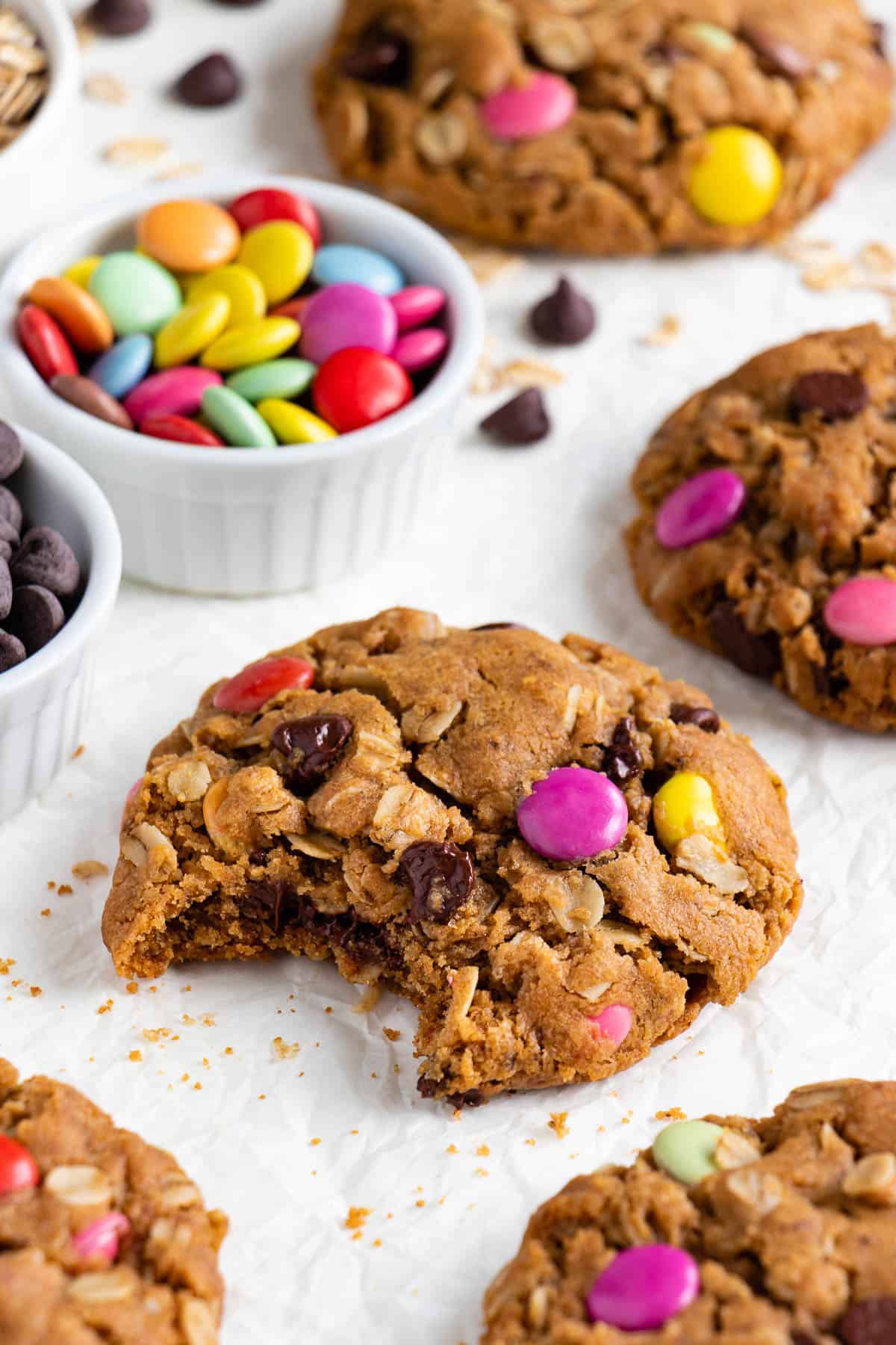 a vegan monster cookie with a bite taken out of it, surrounded by a bowl of vegan m&ms, chocolate chips, and more cookies
