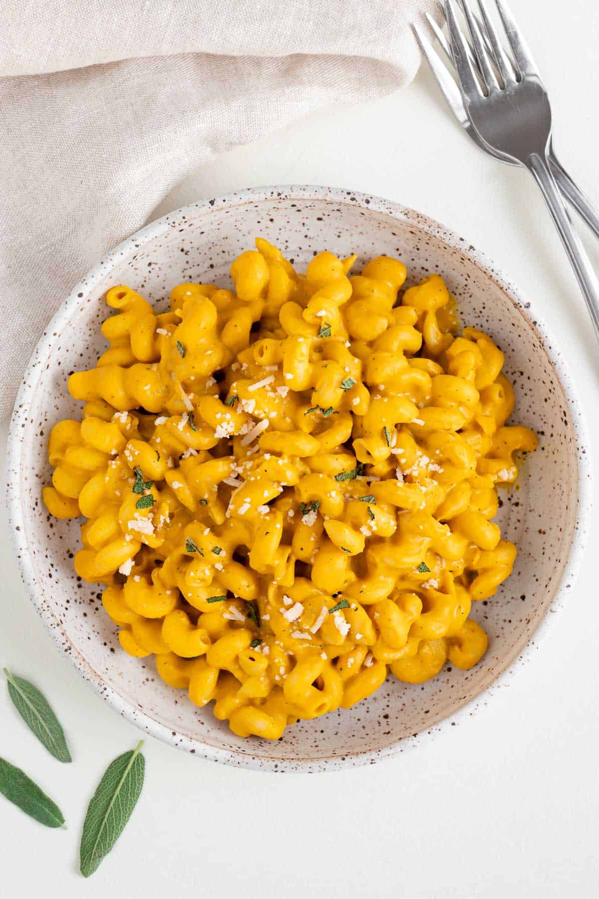 vegan pumpkin pasta inside a ceramic bowl beside two stainless steel forks and fresh sage leaves