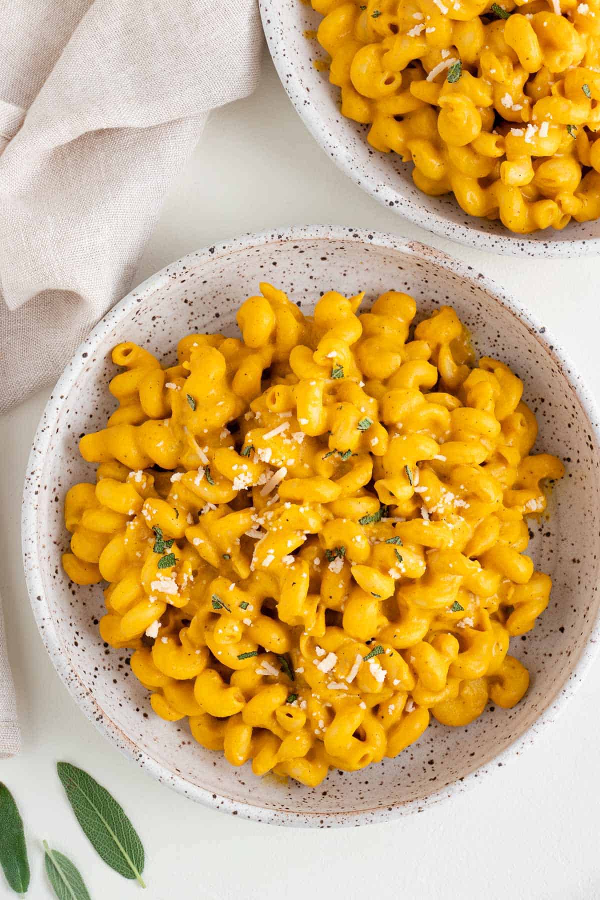 vegan pumpkin pasta inside two ceramic bowls beside a cream colored linen napkin and fresh sage leaves