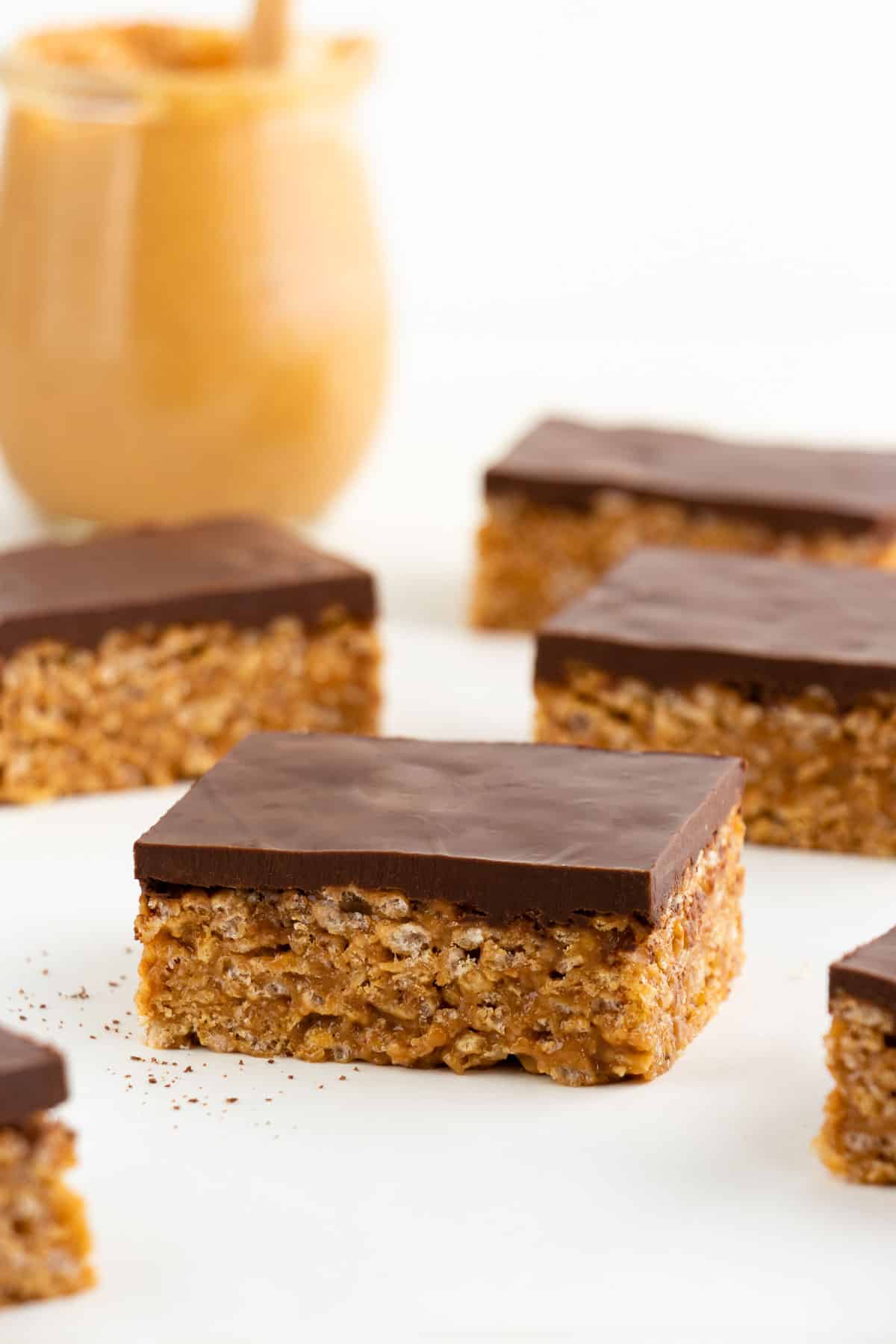 six vegan scotcheroos on a white surface, placed in front of a jar of peanut butter