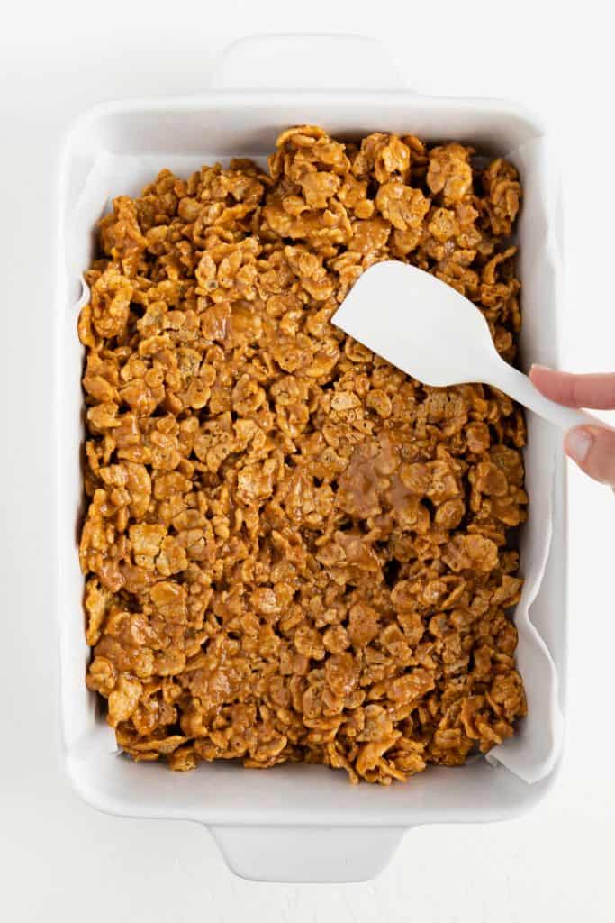 a hand holding a white spatula pressing special k cereal inside a rectangular white baking dish