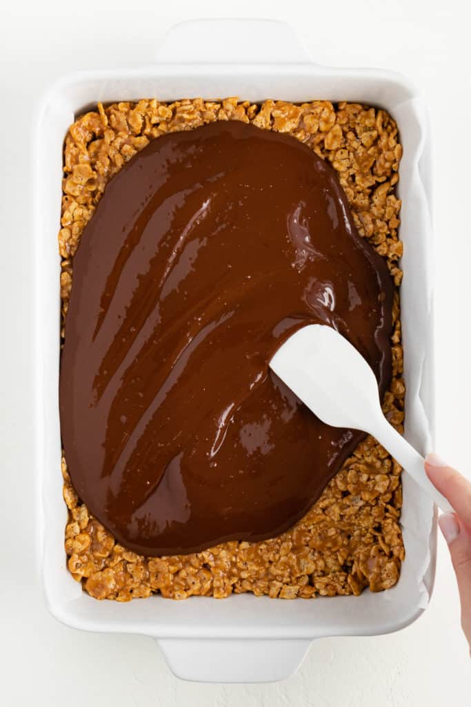 a hand holding a white spatula spreading melted chocolate over the crust of scotcheroo bars