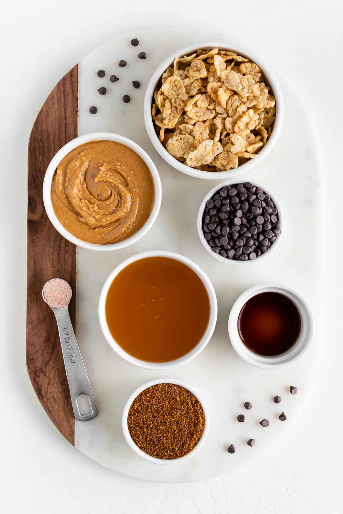 a wood and marble board topped with small bowls of ingredients, including special k cereal, peanut butter, brown rice syrup, chocolate chips, coconut sugar, and vanilla extract