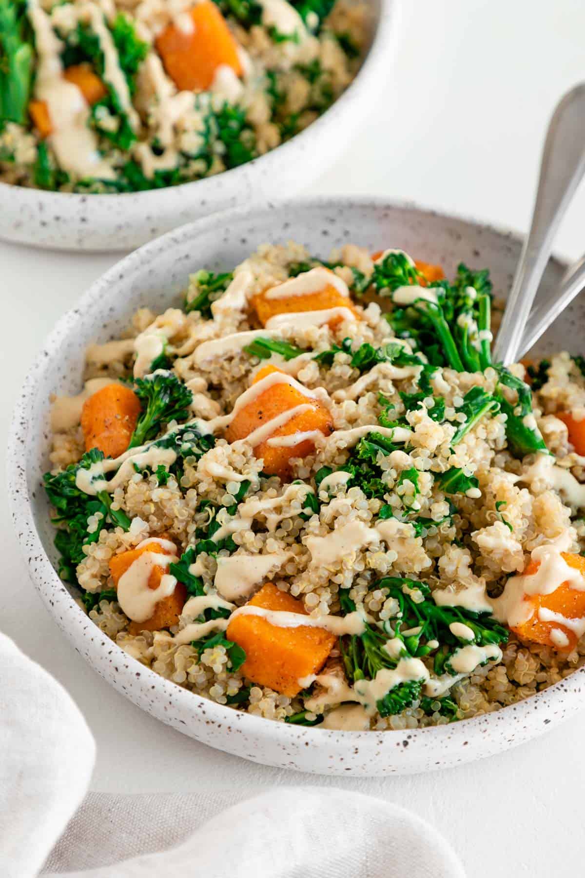 a butternut squash quinoa bowl inside a ceramic dish with two forks, and topped with cashew cream sauce