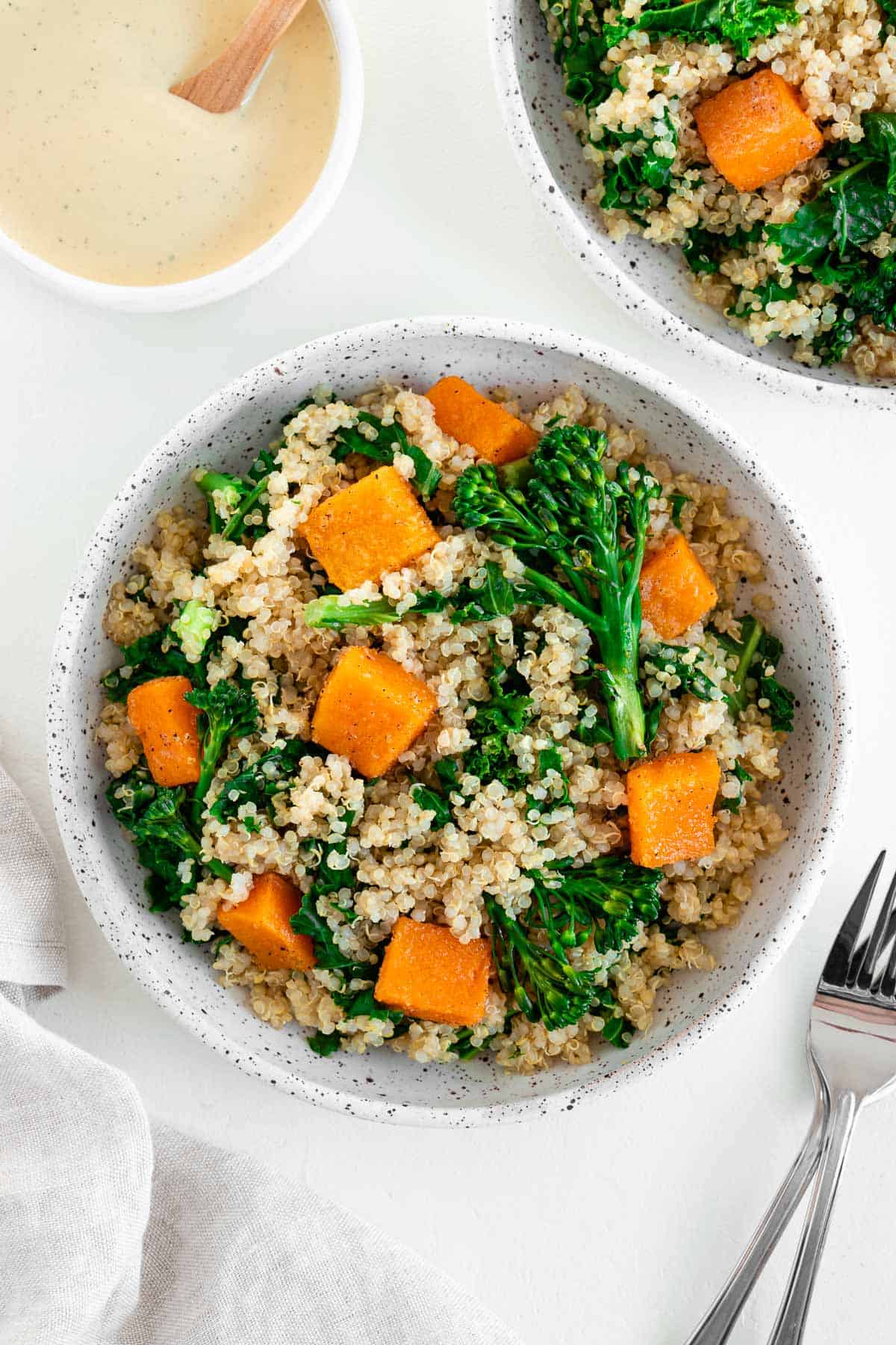 quinoa salad with butternut squash, kale, and broccolini inside two ceramic bowls beside two forks and a bowl of cashew cream sauce