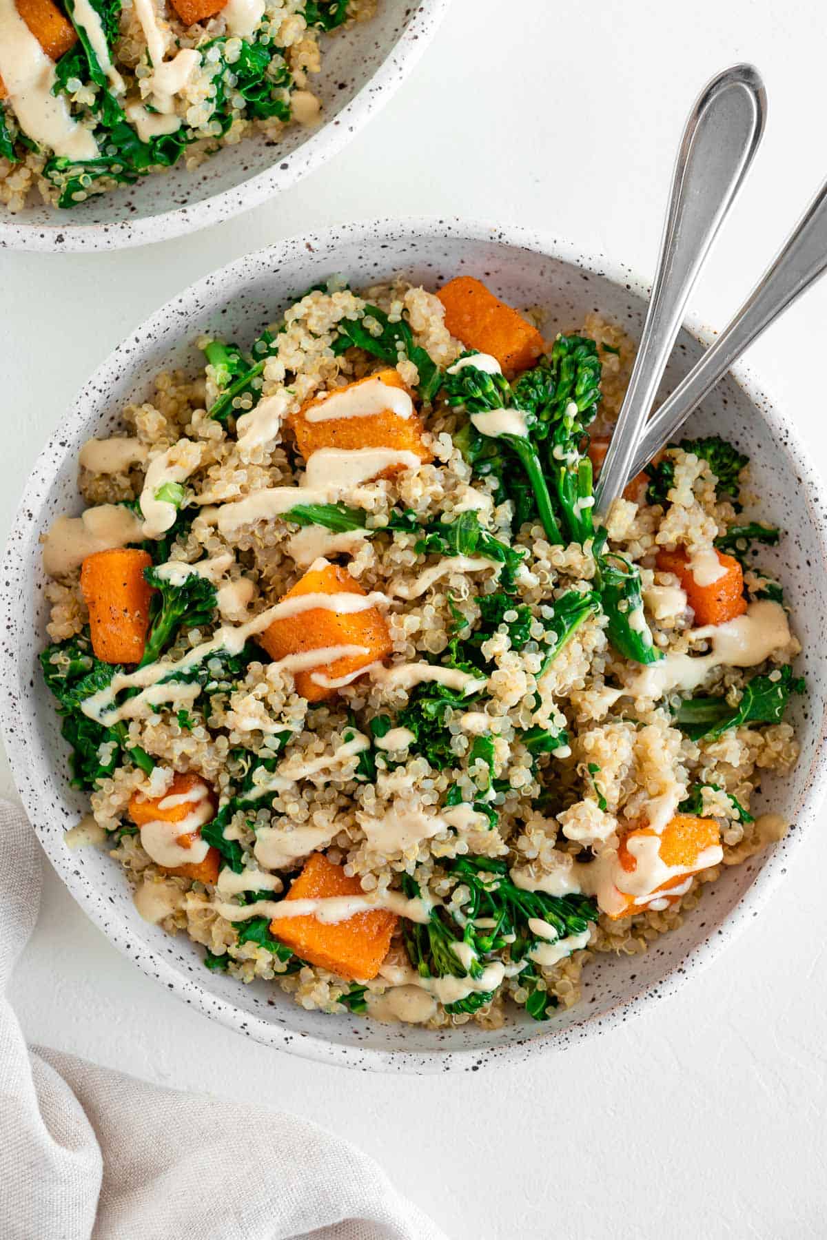 a butternut squash quinoa bowl with kale, broccoli, and cashew cream dressing, placed inside a white ceramic bowl with two forks