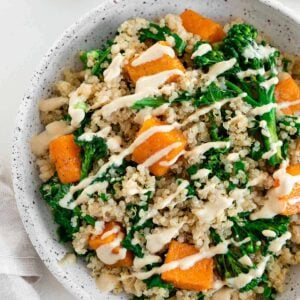 a butternut squash quinoa bowl with kale and broccoli, placed inside a ceramic dish beside a bowl of cashew cream