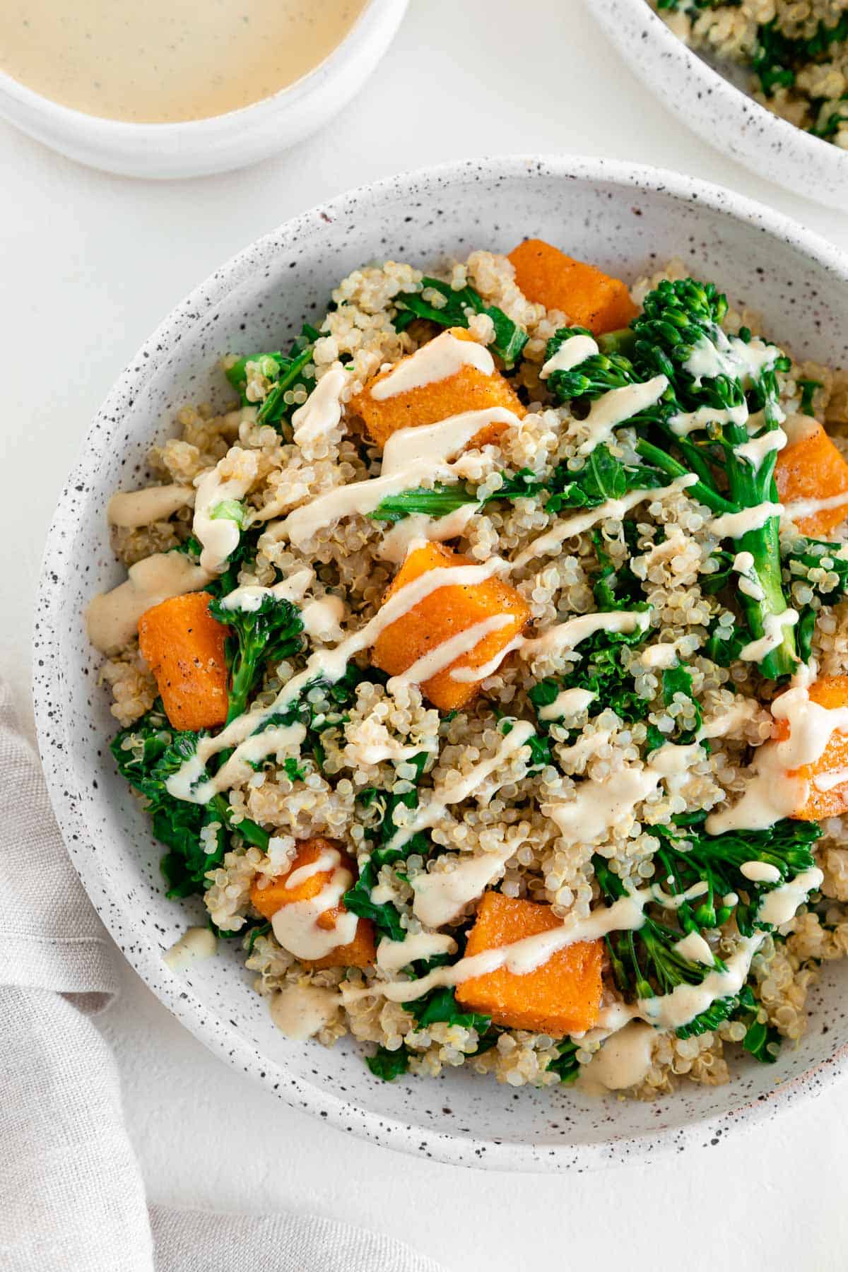 a butternut squash quinoa bowl with kale and broccoli, placed inside a ceramic dish beside a bowl of cashew cream